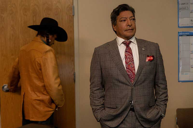 Thomas Rainwater, wearing a brown suit, stands in a room with another character.