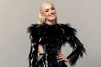 Gwen Stefani, wearing a black feather top, posing for the camera and smiling with her left arm on her hip.