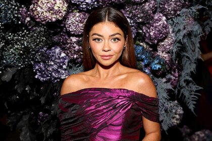 Sarah Hyland poses at an event wearing a purple top with a floral wall in the background.