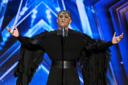 Sheldon Riley, wearing a long black dress with long sleeves with a chain mask, on stage standing in front of a mic.