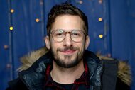 Andy Samberg, wearing a black coat with glasses on, smiles at the camera.