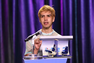 Julio Torres talking behind a podium in front of a purple curtain.