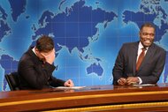 Colin Jost and Michael Che laughing while seated behind a desk.