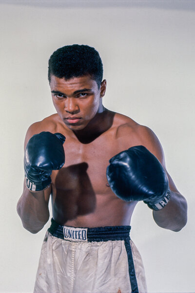 Muhammad Ali posing with his boxing uniform in front of a white backdrop.