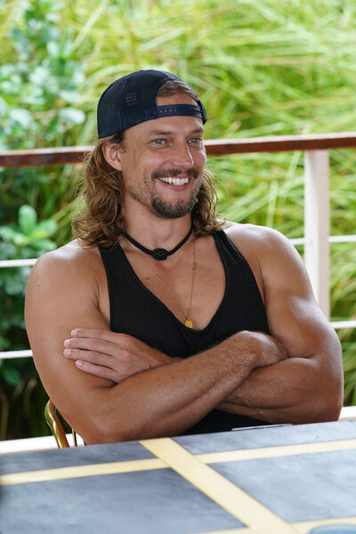 David Genat wearing a black tank top and a baseball cap during Deal or No Deal Island, Season 2