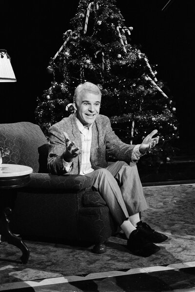 Steve Martin sits in a chair in front of a christmas tree on Saturday Night Live