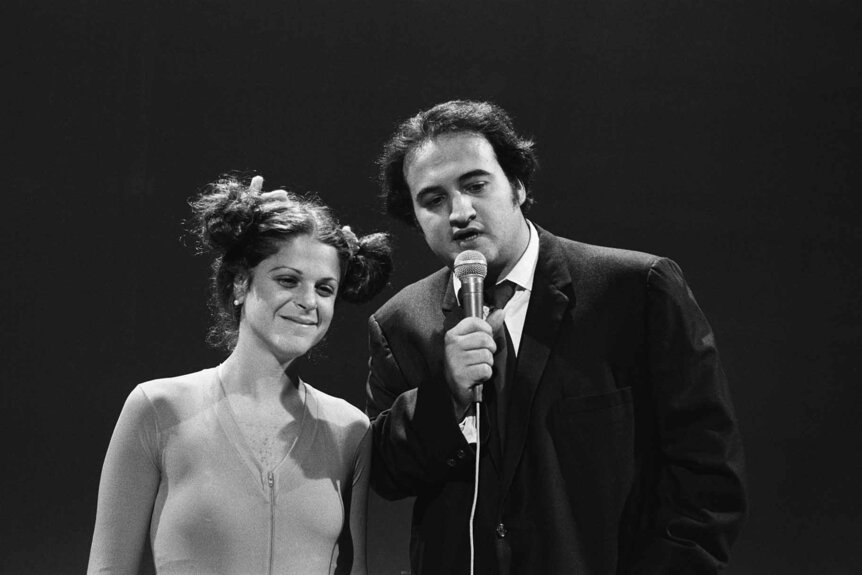 Gilda Radner smiles next to John Belushi who speaks into a mic on Saturday Night Live Season 1 Episode 23.