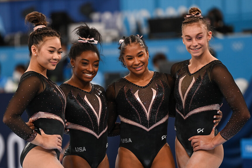 Sunisa Lee, Simone Biles, Jordan Chiles and Grace McCallum smile together in their gymnastic leotards.