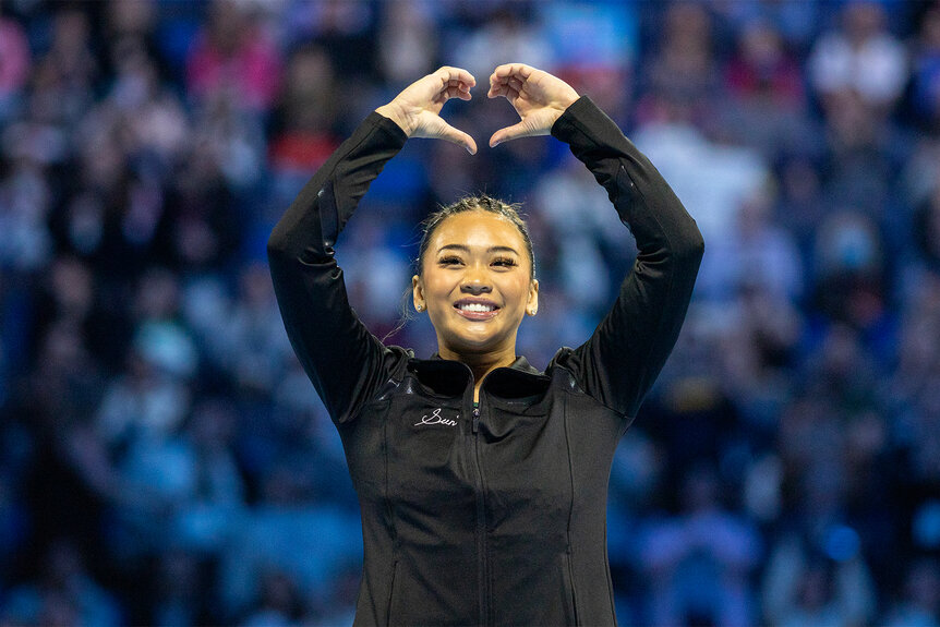 Suni Lee makes a heart with her hands to the crowd at a gymnastics meet
