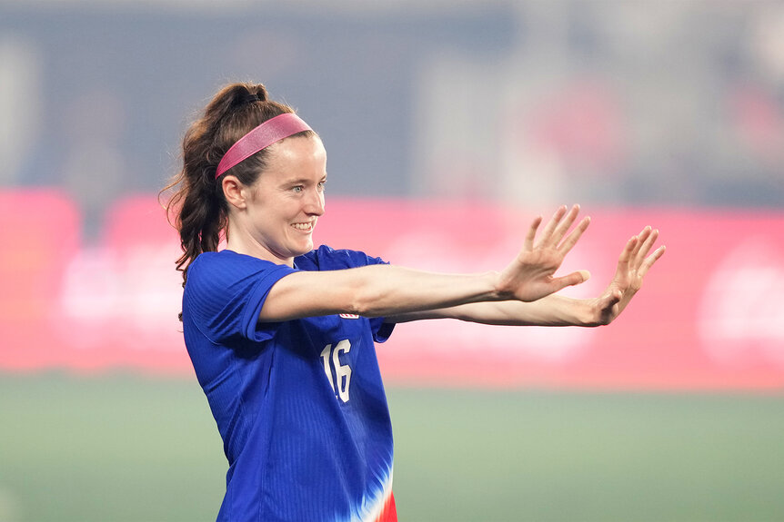 Rose Lavelle reaches her arms out and smiles on the soccer pitch