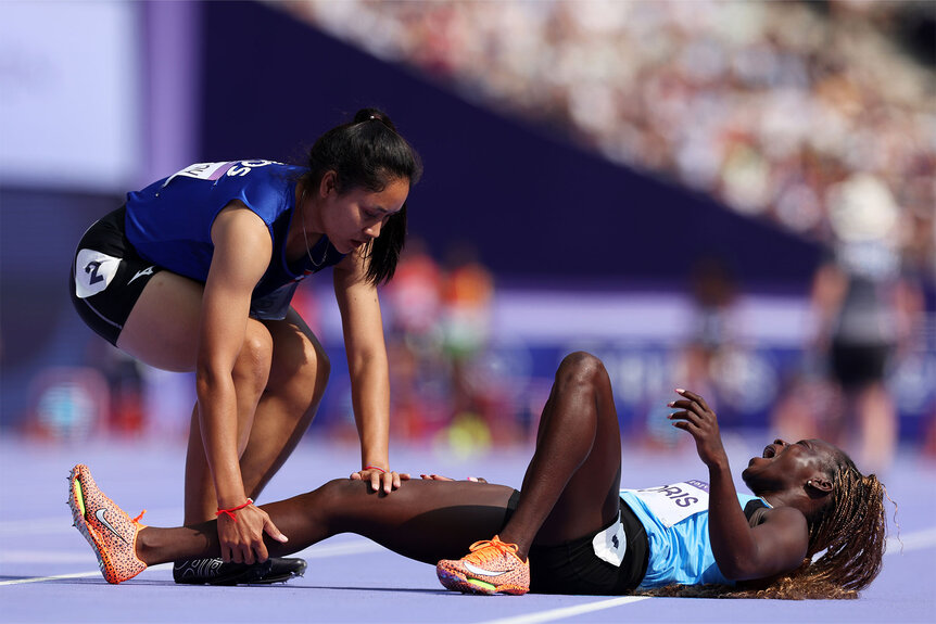 Lucia Moris of Team South Sudan lies injured on the ground as Silina Pha Aphay of Team Lao checks her condition during the Women's 100m Preliminary Round on day seven of the Olympic Games Paris 2024