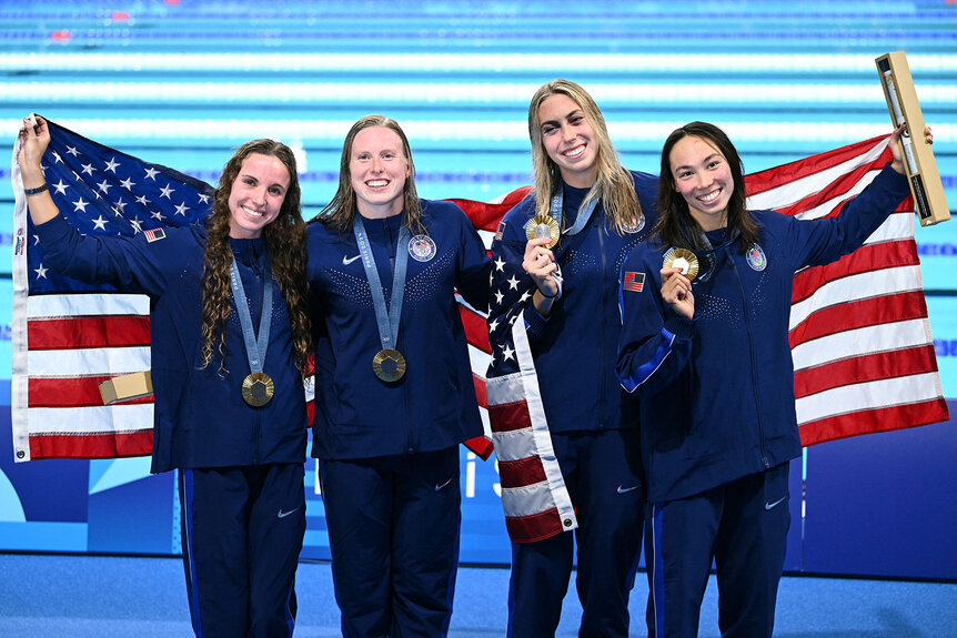 Regan Smith, Lilly King, Gretchen Walsh and Torri Huske pose with their medals and American flags in front of a pool at the Paris 2024 Olympics