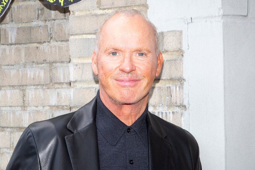 Michael Keaton smiles while wearing a leather blazer and grey shirt