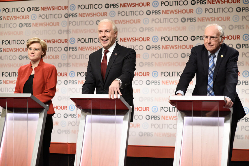 Kate McKinnon as Elizabeth Warren, Jason Sudeikis as Joe Biden, and Larry David as Bernie Sanders standing behind debate podiums