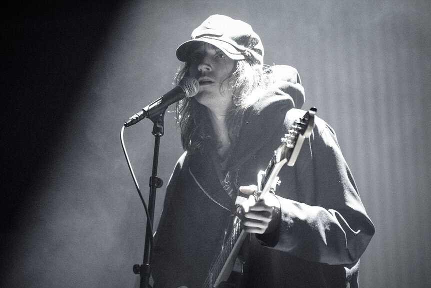 Mk.gee singing and playing guitar during an onstage performance.