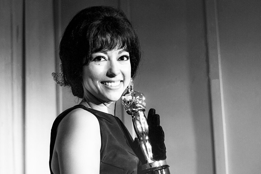 Rita Moreno holds up her Oscar in a dark dress smiling