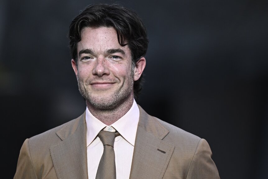 John Mulaney smiles in beige suit.