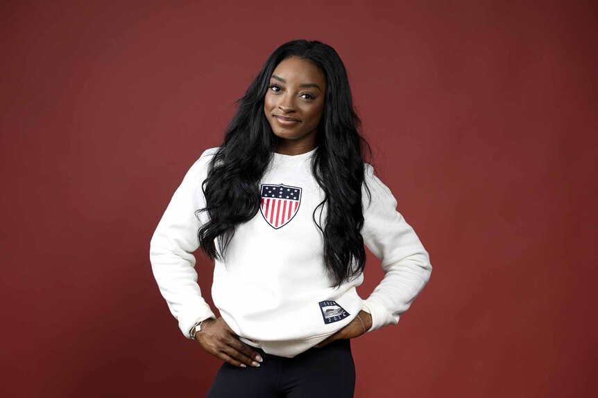 Simone Biles wears a white sweatshirt in front of a red backdrop.