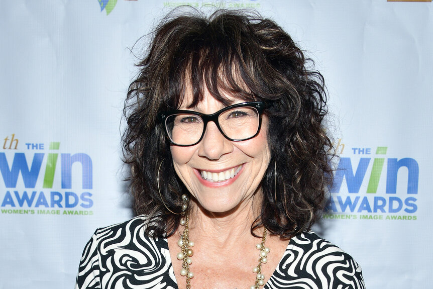 Mindy Sterling smiles on the red carpet in a black and white shirt