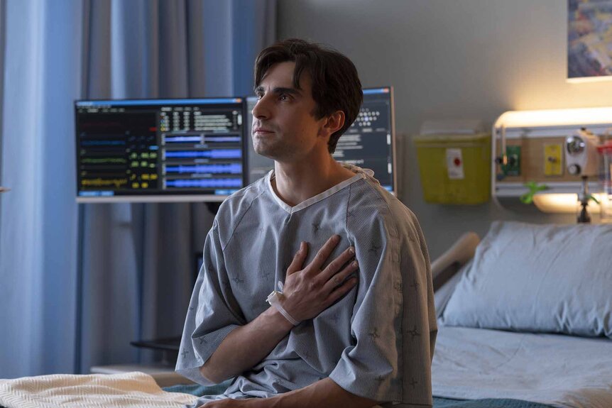 Roman (John Doe) (Alex Ozerov-Meyer) wears a hospital gown and holds his chest in Brilliant Minds Episode 107.