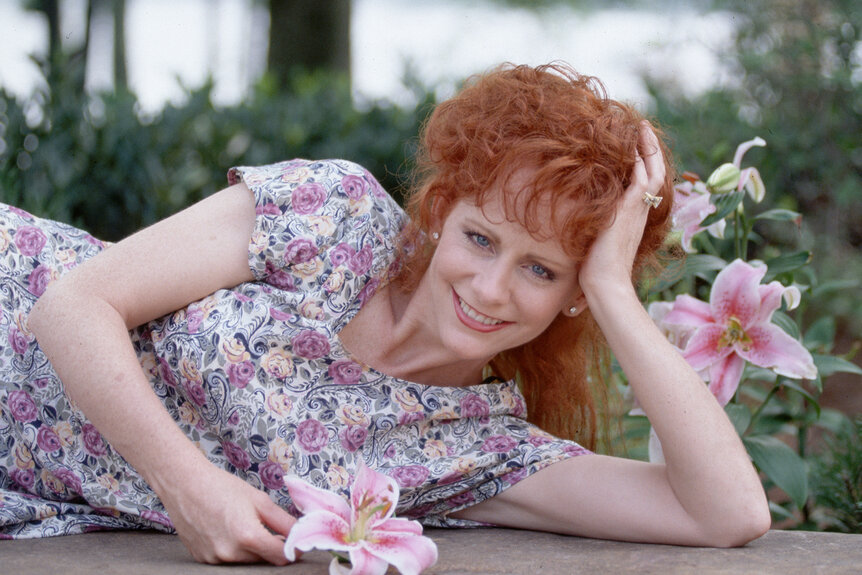 Reba Mcentire lays on her side in a floral dress while holding a flower