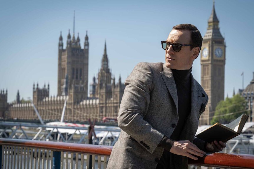 George Woodhouse (Michael Fassbender) leans against a railing while holding a book in Black Bag (2025).