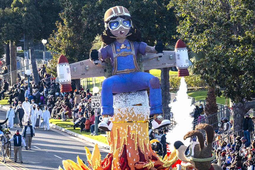 A floral arrangement parade float in the shape of a woman with a jetpack is walked on a road.