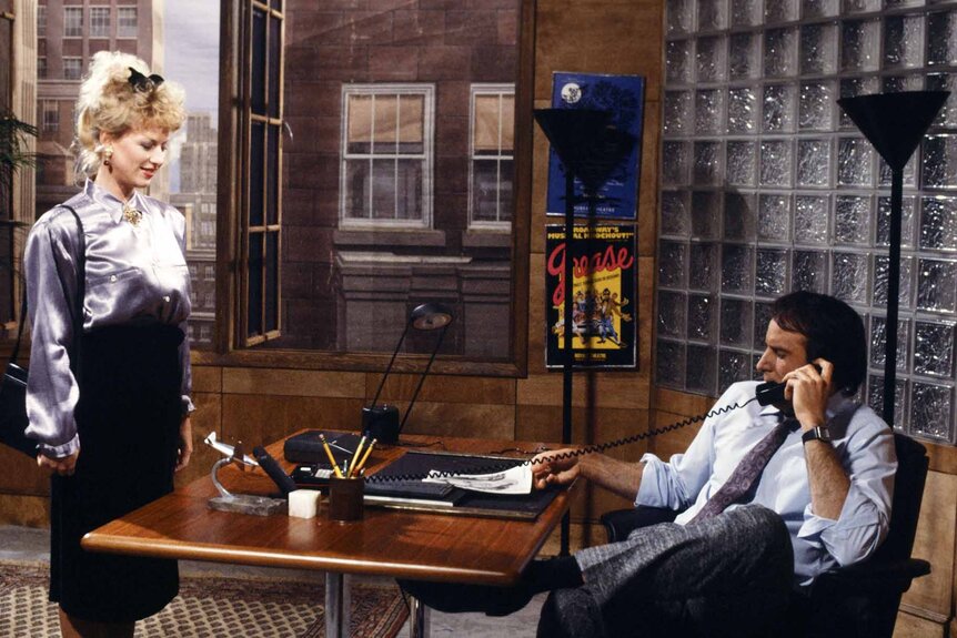 Actress (Victoria Jackson) stands next to Agent (Kevin Nealon) who speaks on the phone at his desk on SNL.