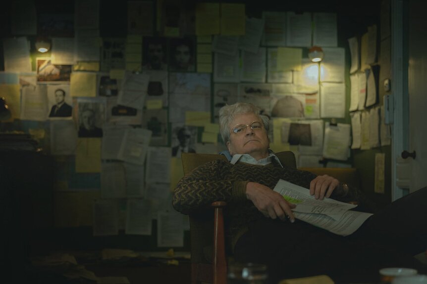 Jim Swire (Colin Firth) reclines in a chair while holding a newspaper in Lockerbie: A Search for Truth Episode 103.