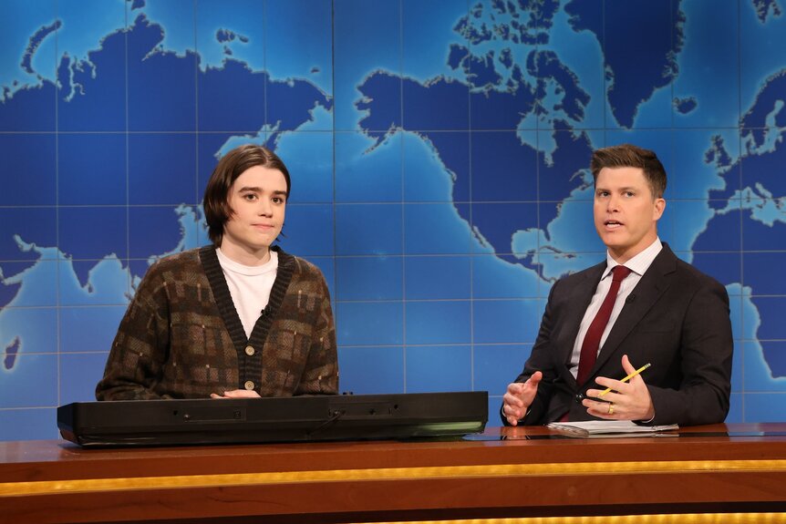 Jane Wickline and Colin Jost sitting behind a desk in front of a map.