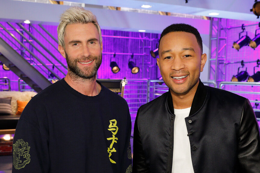 Adam Levine and John Legend smile together with a illuminated purple light in the background.